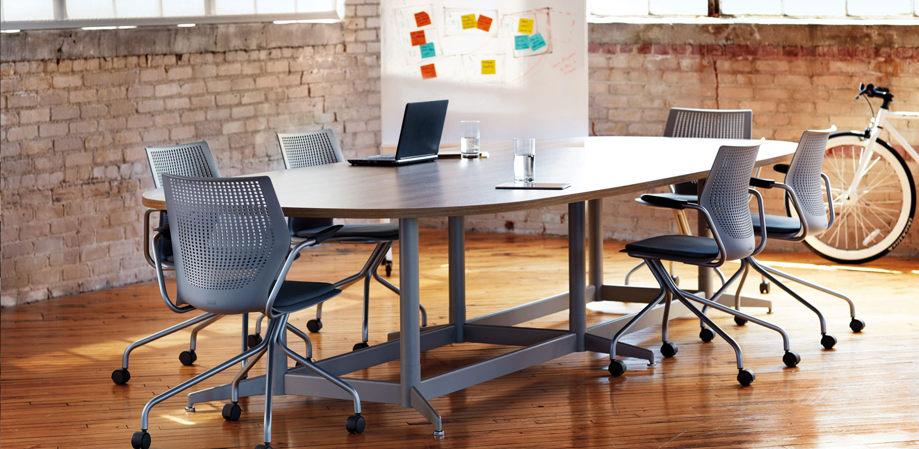 Modern board room with brick walls and wood floors.  There is a white board with sticky notes in the background, and a bike leaning against the wall.