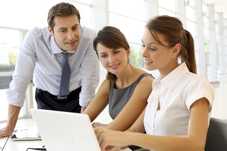 Two women sitting at a table, looking at a laptop, while a young man leans over listening.