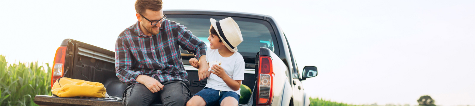 Young father in plaid shirt and jeans, with his son in blue shorts and a white t-shirt wearing a fedora.  They are smiling and laughing while sitting in the bed of a pickup, parked in field.
